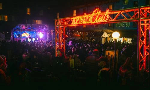 Nighttime scene at a music festival with a crowd and 'Apres Club' sign.