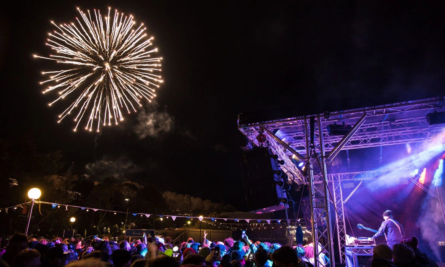 Fireworks exploding in the night sky above a concert stage with a DJ.
